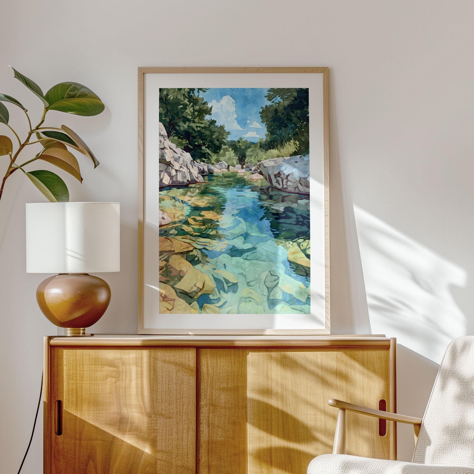 Framed watercolor-style painting of a rocky river with turquoise and green water, surrounded by trees and a bright blue sky, displayed on a light wooden cabinet. A round brown lamp with a white shade sits beside the artwork, with a plant and modern armchair completing the cozy, sunlit interior.