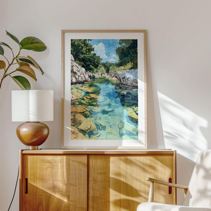 Framed watercolor-style painting of a rocky river with turquoise and green water, surrounded by trees and a bright blue sky, displayed on a light wooden cabinet. A round brown lamp with a white shade sits beside the artwork, with a plant and modern armchair completing the cozy, sunlit interior.