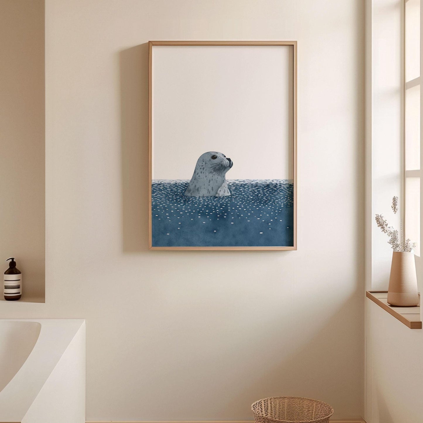 Framed minimalist art print of a gray spotted seal rising from dark blue water with scattered white ripples, hung on a beige bathroom wall. The serene scene is styled with soft natural light, a woven basket, a white vase with dried stems on the windowsill, and a modern tub, creating a calm, spa-like atmosphere.