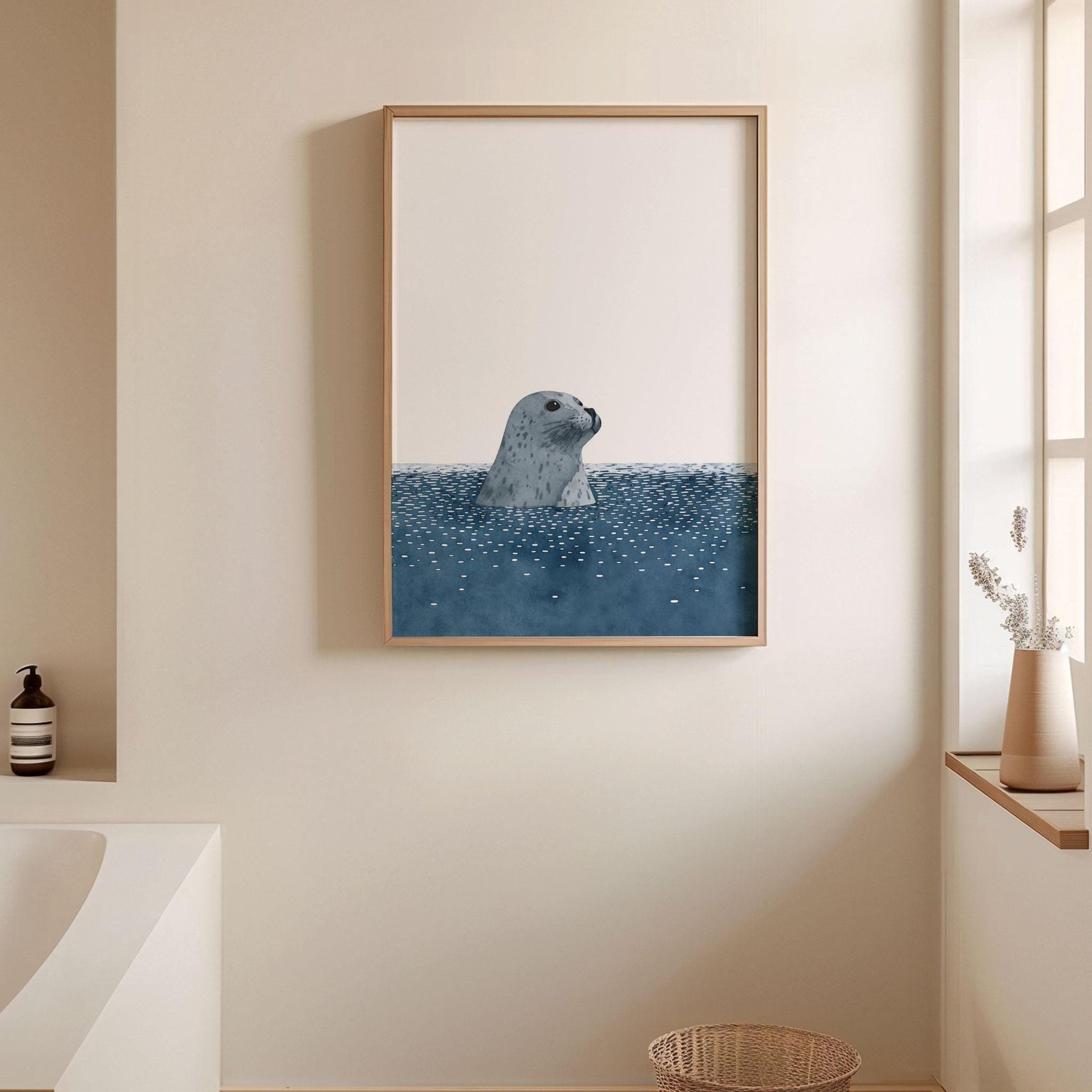 Framed minimalist art print of a gray spotted seal rising from dark blue water with scattered white ripples, hung on a beige bathroom wall. The serene scene is styled with soft natural light, a woven basket, a white vase with dried stems on the windowsill, and a modern tub, creating a calm, spa-like atmosphere.