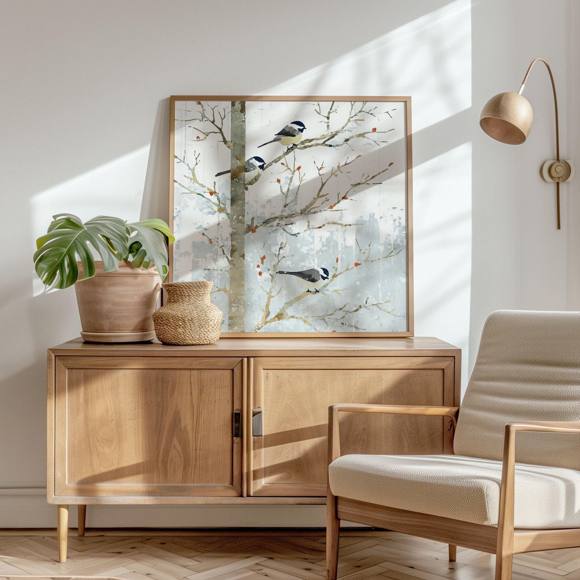 Framed watercolor-style print of three chickadees on bare winter branches, propped on a light wood cabinet. Soft blue-gray snowy background contrasts with the warm wood tones. A potted monstera, woven vase, cream armchair, and brass wall lamp complete the bright, modern, sunlit room.