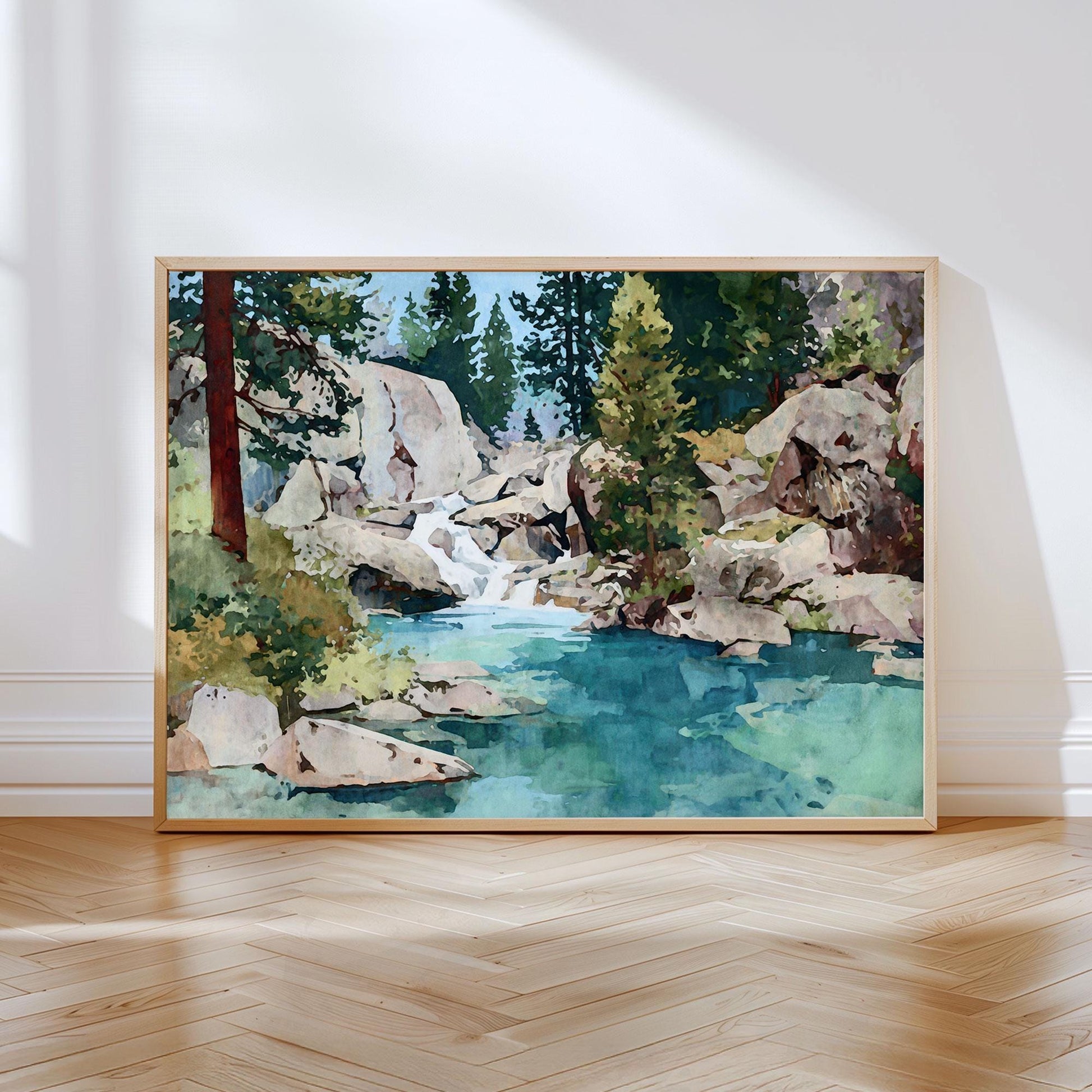 Watercolor landscape art print depicting a serene mountain stream flowing over rocks into a turquoise pool, surrounded by evergreen trees and granite boulders under a clear blue sky. The artwork rests in a light wood frame on a herringbone floor, leaning against a white wall with soft natural light casting gentle shadows.