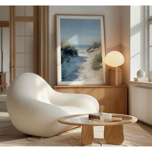 Modern living room with a coastal art print of a sandy beach path framed and leaning against the wall on a wooden sideboard, beside a warm glowing lamp. A textured white lounge chair and a round wooden coffee table create a cozy, minimalist setting.