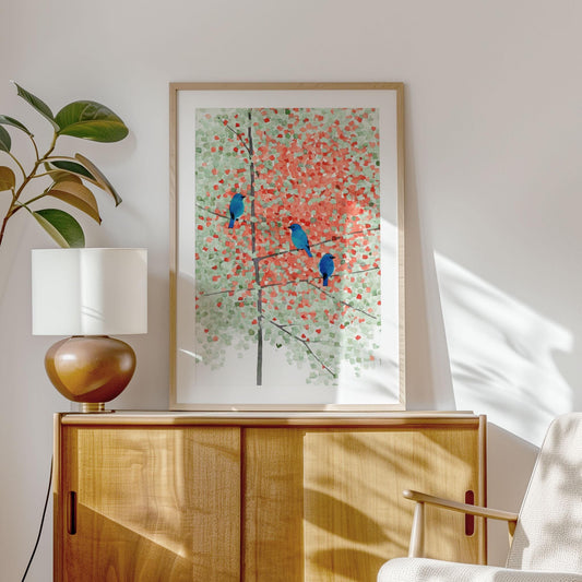 Framed art print of three bright bluebirds perched on maple branches with soft green and red leaves, painted in a mosaic watercolor style. Displayed above a mid-century wood credenza with a ceramic lamp and indoor plant, in a sunlit modern living room with warm natural tones.