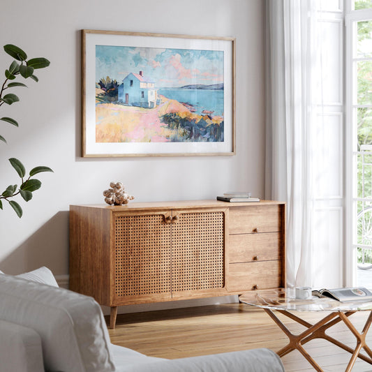 Framed watercolor painting of a coastal scene with a blue house on a bluff overlooking the ocean, displayed above a wooden cabinet with woven rattan doors. The room features light walls, sheer white curtains, a glass coffee table with books and cups, and a potted plant, creating a bright and airy coastal-inspired living space.