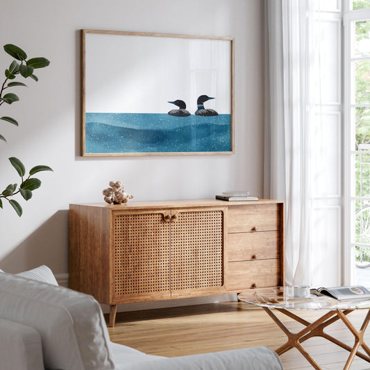 Framed minimalist loon artwork hangs above a mid-century modern wooden sideboard with woven cabinet doors. The print shows two black-and-white loons floating on deep blue speckled water against a white background. The scene is styled in a bright living room with white walls, sheer curtains, a leafy plant, and a glass coffee table, creating a calm, natural atmosphere.