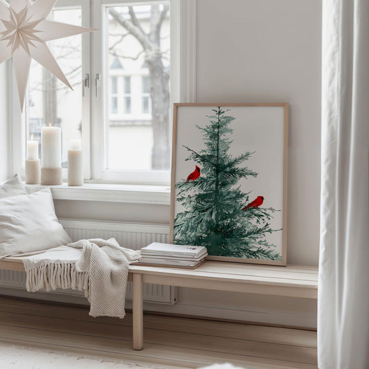 Cozy Scandinavian-style room with a framed watercolor print of a pine tree and two red cardinals leaning against the wall on a wooden bench, next to stacked books, a white throw blanket, and large candles by a window.