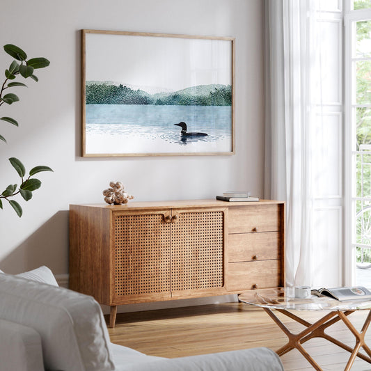 Framed art print of a loon gliding across a calm blue lake with green forested hills in the background, displayed above a mid-century wooden sideboard with woven cabinet doors in a bright, airy living room with natural light, white walls, and minimalist decor.