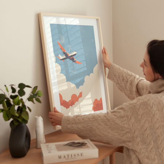 Woman in a cozy beige sweater places a framed art print of an airplane with orange wings flying through stylized clouds against a blue sky on a wooden table; nearby are a black vase with greenery, a sculptural white object, and a Matisse art book.