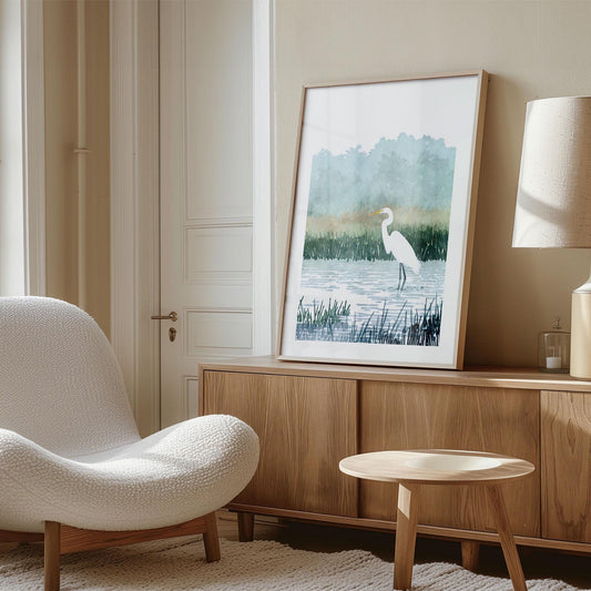 Framed watercolor-style print of a white egret standing in a marsh, leaning against the wall on top of a wooden sideboard in a modern living room. The room features a textured white lounge chair, round wooden coffee table, beige walls, and a neutral-toned lamp, creating a calm and minimalist aesthetic.