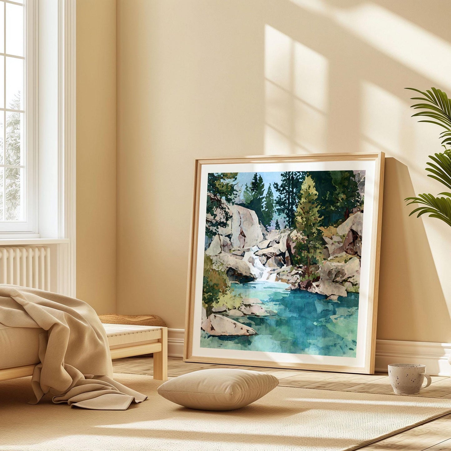 Framed watercolor landscape art print of a clear turquoise mountain stream surrounded by pine trees and granite boulders, leaning against a beige wall in a sunlit minimalist bedroom. The room features soft neutral tones, a low wood bed with a beige blanket, a floor cushion, and a ceramic mug on a woven rug, creating a calm and cozy natural atmosphere.