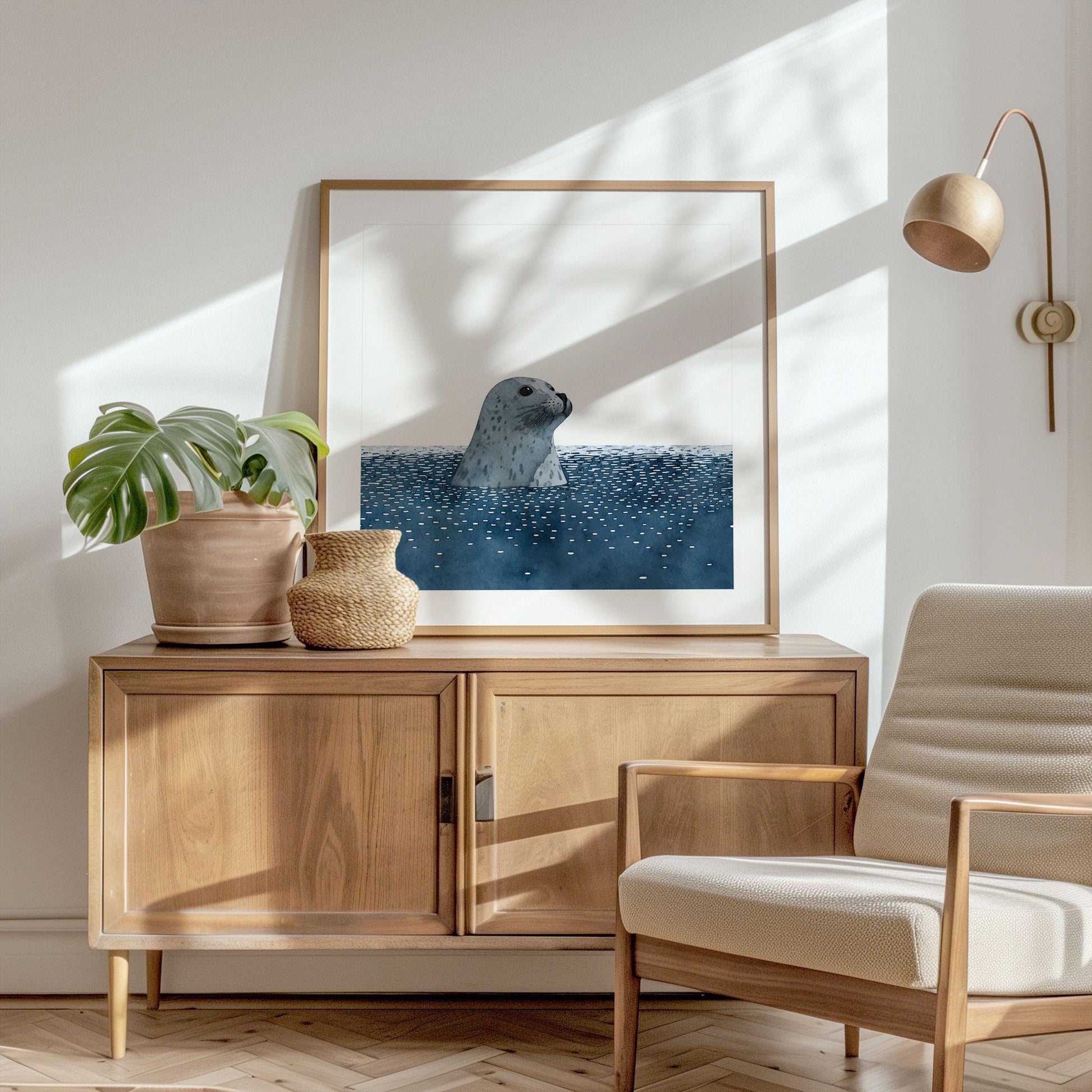 Framed art print of a gray spotted seal emerging from deep blue water, displayed on a wooden sideboard in a sunlit living room. The minimalist coastal artwork is styled with a potted monstera plant, a woven vase, and a neutral armchair, with soft natural light and shadows creating a calm, modern atmosphere.