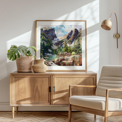 Framed mountain landscape painting displayed on a mid-century wood sideboard in a sunlit room. The artwork features rugged peaks, pine trees, and a turquoise river rendered in a modern impressionist style. A potted monstera plant and woven vase sit beside the frame, with a cream upholstered armchair and brass wall lamp completing the warm, minimalist interior.
