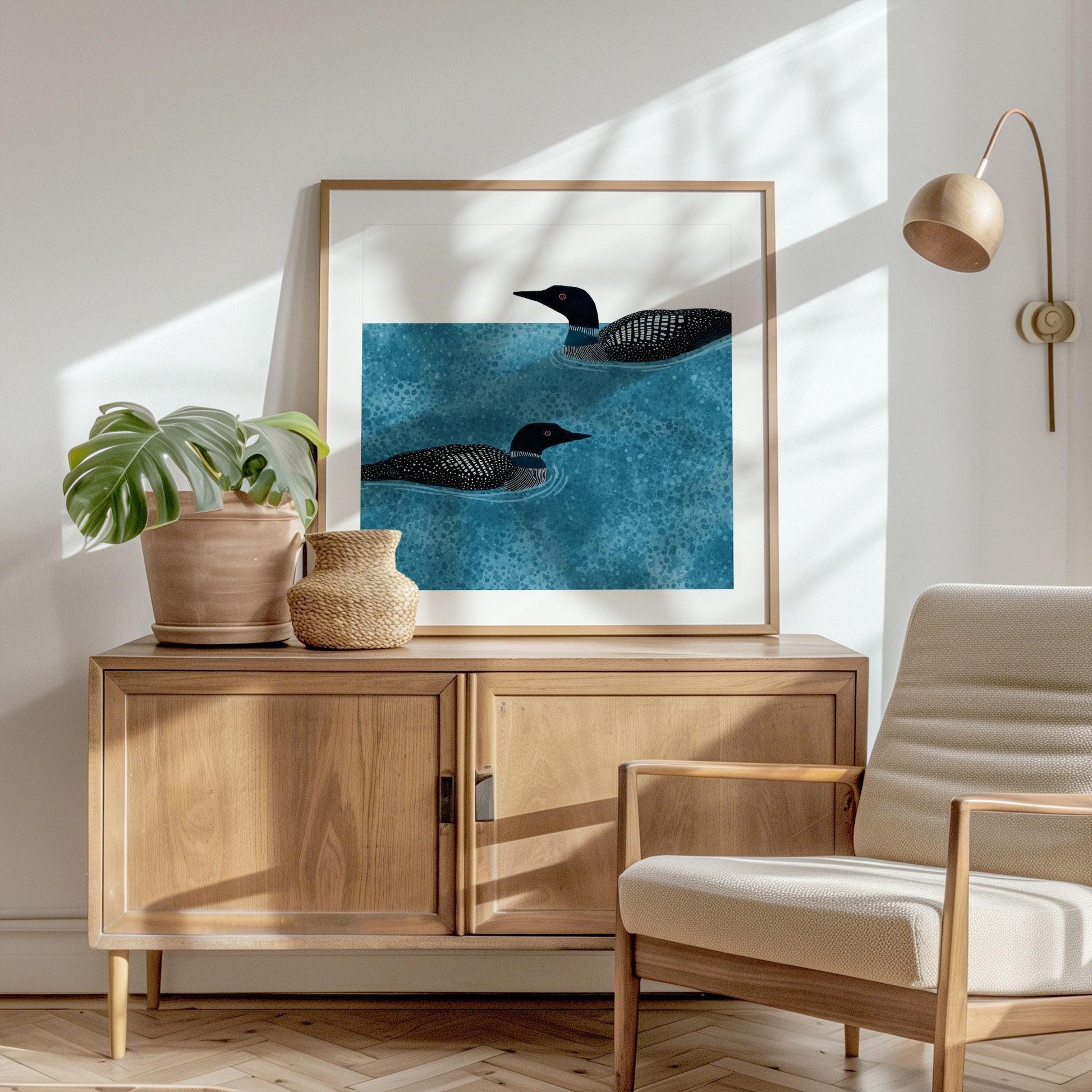 Framed loon illustration with two black and white loons swimming in textured blue water, displayed on a wooden cabinet. The scene includes a potted monstera plant, woven vase, and a light wood armchair with cream upholstery. Sunlight streams through, casting soft shadows on the white wall, complemented by a modern wall-mounted lamp.