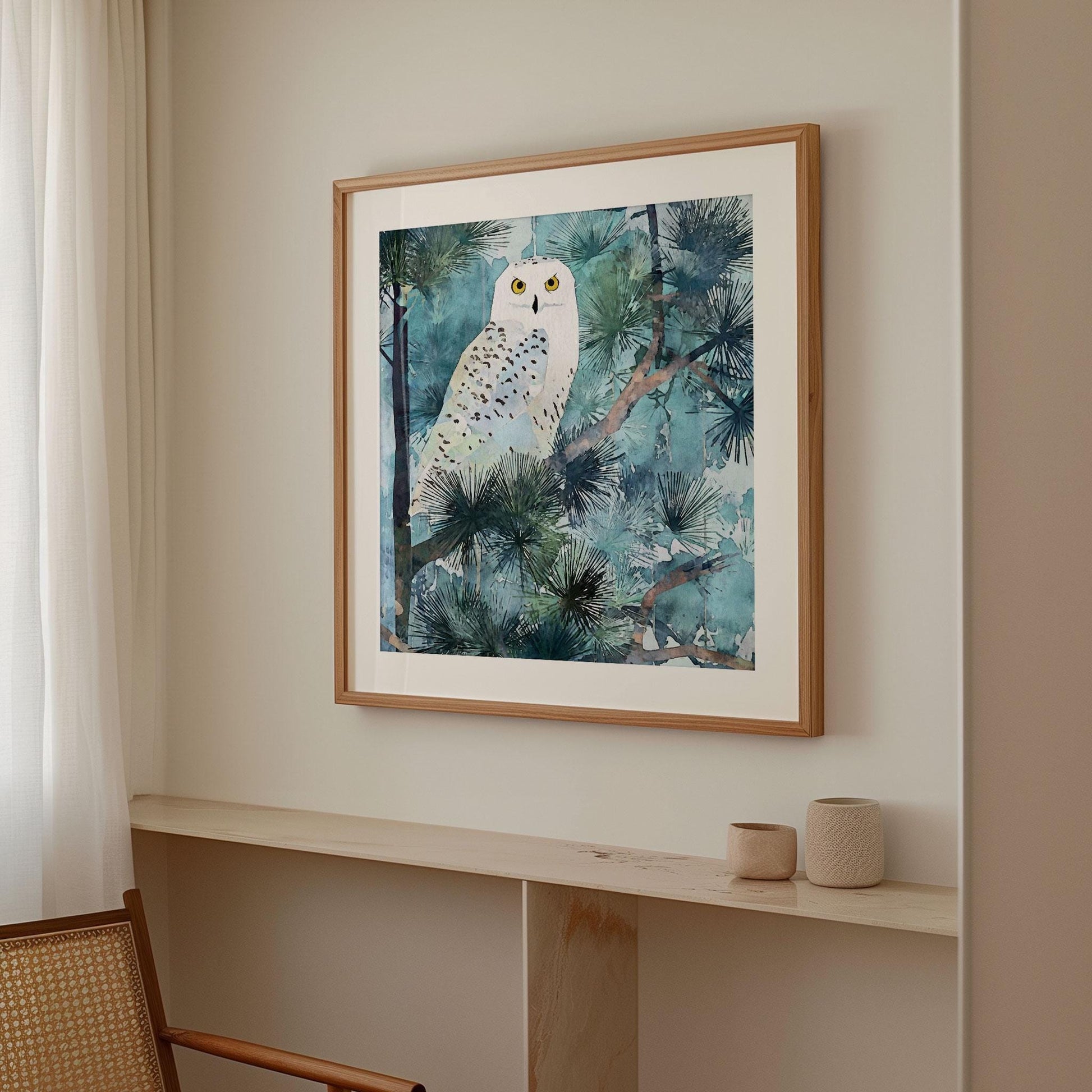 Framed watercolor-style art print of a snowy owl with bright yellow eyes perched among pine branches, in cool blue and green tones. The artwork hangs on a light neutral wall above a minimalist wooden ledge with two small ceramic vessels, next to a woven chair and soft white curtains.