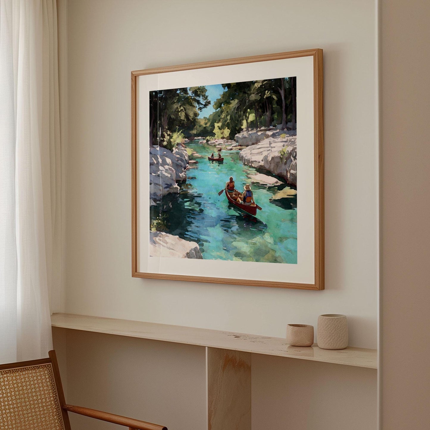 Framed painting of two people paddling a red canoe on a turquoise river with rocky cliffs and trees, hanging on a neutral wall in a softly lit modern room with light wood accents and minimal decor.