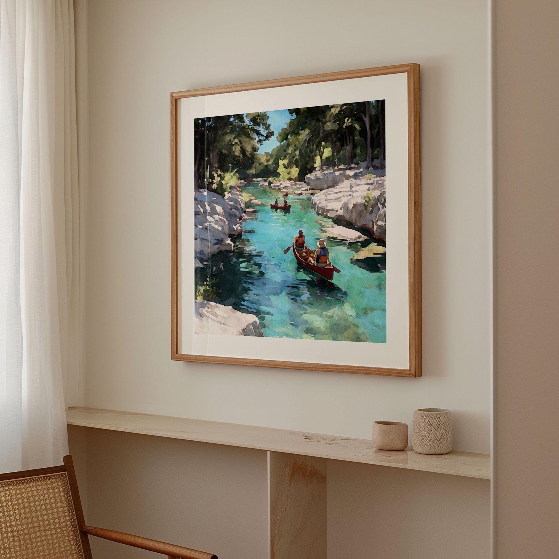 Framed painting of two people paddling a red canoe on a turquoise river with rocky cliffs and trees, hanging on a neutral wall in a softly lit modern room with light wood accents and minimal decor.