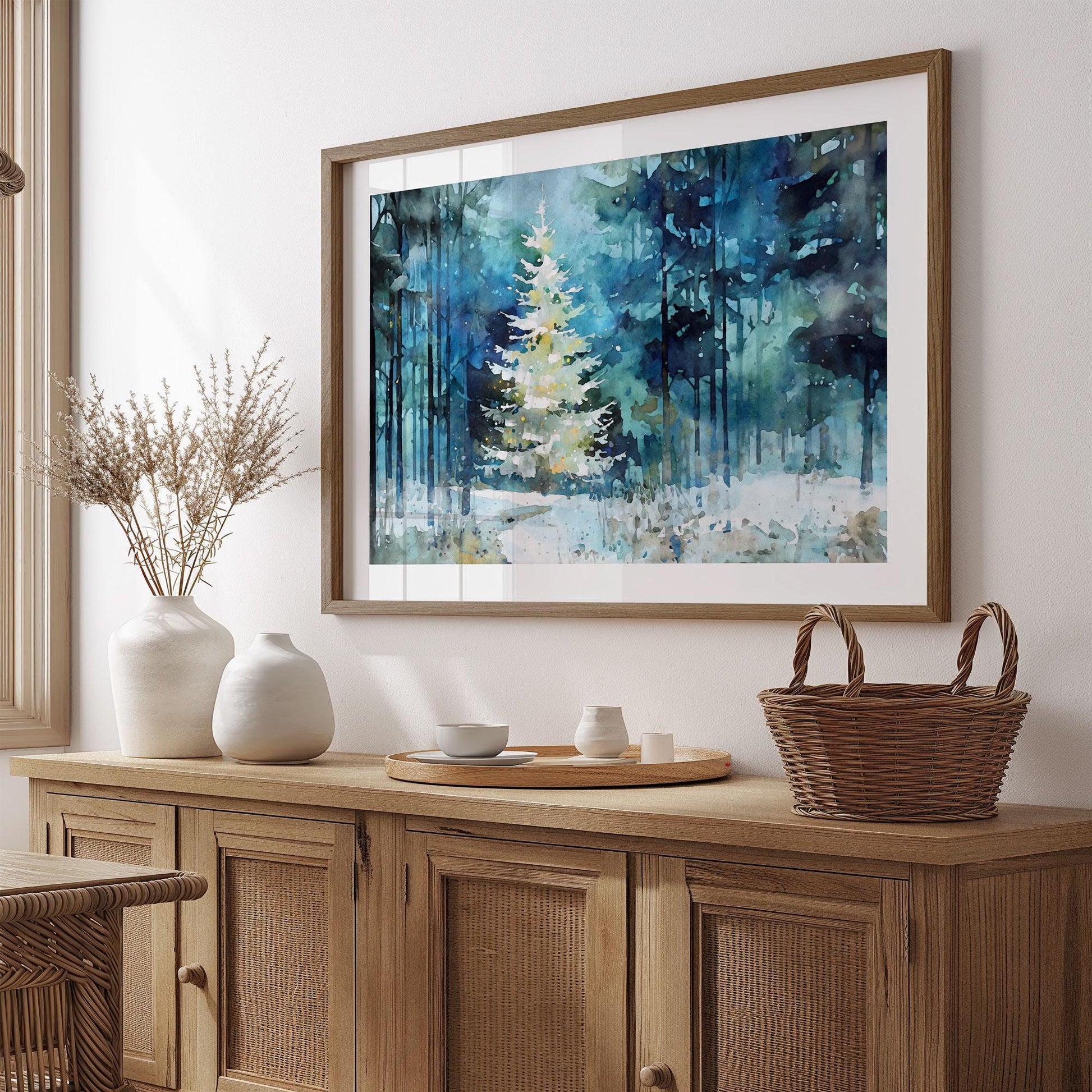 Framed watercolor art of a glowing white Christmas tree in a snowy forest, hung above a natural wood sideboard. Soft blues and greens fill the background with tall trees. The scene is styled with neutral décor—ceramic vases, a tray with small vessels, and a woven basket—creating a warm, rustic holiday setting.