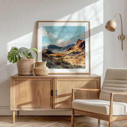 Framed painting of a desert canyon with sunlit red rock cliffs, golden hills, and distant blue mountains under a bright sky with fluffy clouds, displayed on a wooden sideboard beside a potted plant and woven vase in a sunlit modern living space with a neutral armchair.