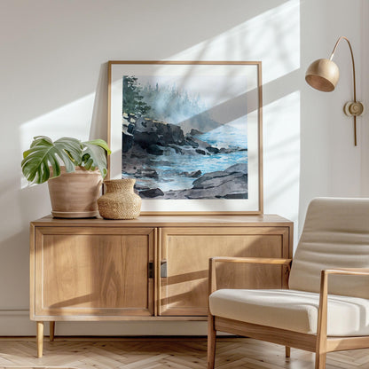 Framed horizontal art print displayed on a wooden cabinet in a sunlit living room. The watercolor-style artwork depicts a misty Lake Superior shoreline with dark rocky cliffs, calm blue water, and soft fog drifting across a distant evergreen forest. Warm natural light, indoor plants, and mid-century–style furnishings create a serene, modern interior setting that complements the artwork’s muted blues, grays, and greens.