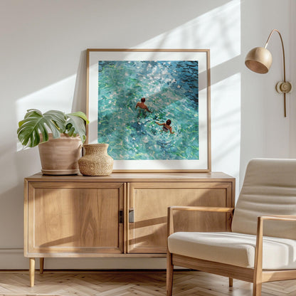 Framed square painting of two swimmers seen from above in sunlit turquoise water, with dappled light and rippling waves, displayed on a wooden sideboard beside potted plants in a bright, modern room with natural textures.