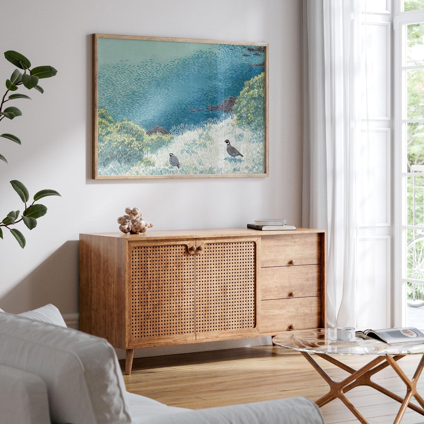 Framed California quail art print displayed in a sunlit living room above a mid-century wood and rattan sideboard. The artwork features two quail in a stylized coastal meadow with lush grasses and a deep blue ocean beyond, creating a tranquil, nature-inspired focal point in the airy, minimalist space.