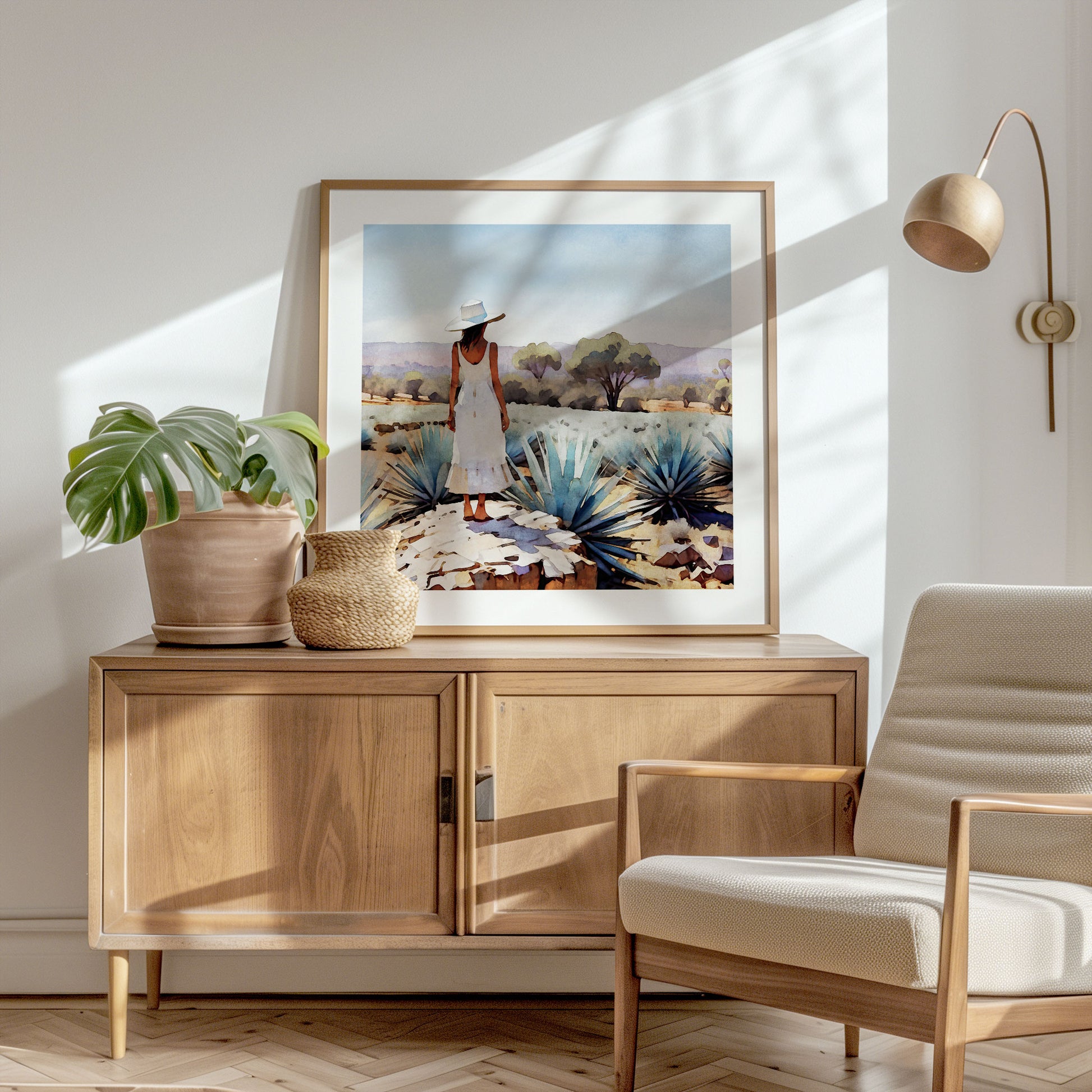 A sunlit corner with a wooden sideboard features a framed artwork of a woman in an agave field, alongside a potted monstera plant and a woven basket.