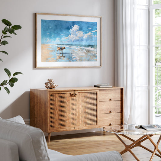 Framed watercolor-style beagle beach painting displayed above a mid-century wood sideboard in a bright, airy living room. The artwork shows a tricolor beagle walking along the shoreline with reflections in wet sand, beneath a blue sky with soft clouds, styled with neutral decor, plants, and natural light.