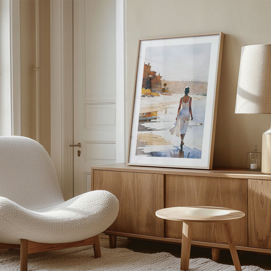 A framed watercolor painting of a woman in a white dress walking along the beach leans against the wall on a wooden sideboard. 