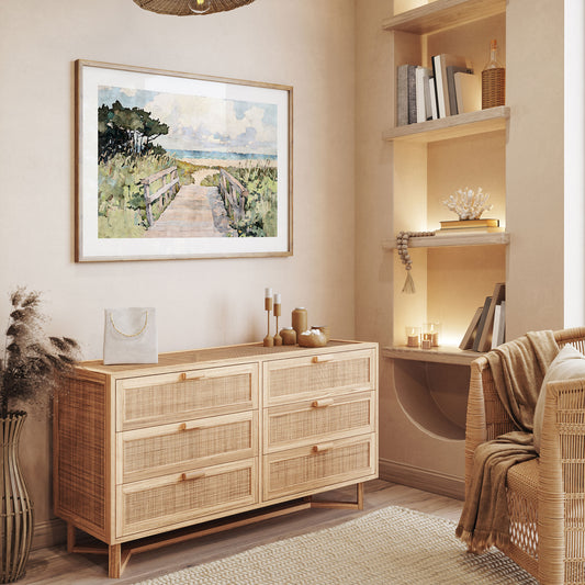Framed beach boardwalk art print displayed in a warm, coastal-inspired room with natural wood and rattan furniture. The artwork features a watercolor-style path leading through seaside dunes toward the ocean, with soft clouds and a calm blue horizon. Decor elements like a woven chair, neutral candles, and coral accents complement the serene coastal aesthetic.