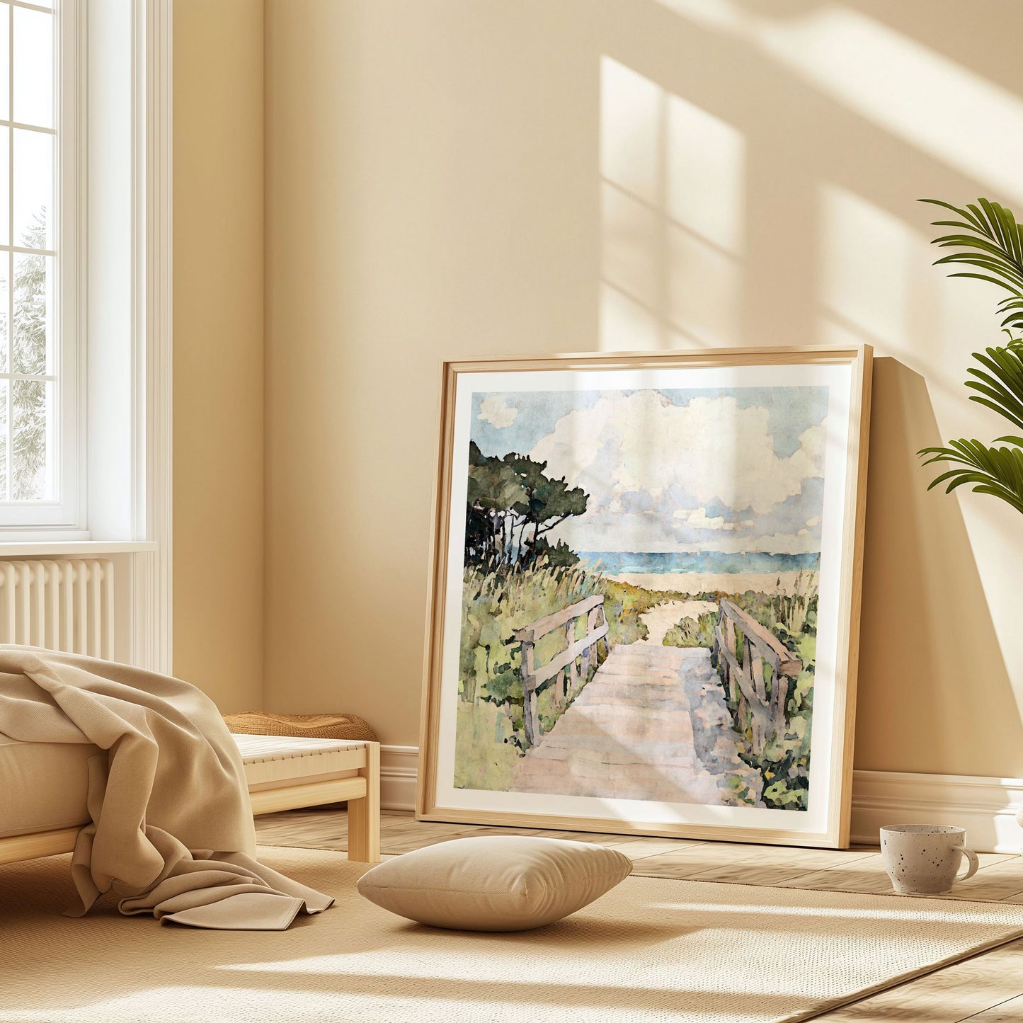 Large beach boardwalk art print in a minimalist, sunlit room with pale wood tones and neutral decor. The painting features a watercolor path leading to the ocean, flanked by grasses and trees under a cloudy sky. The print leans casually against the wall near a floor cushion, cozy blanket, and coffee mug, creating a relaxed coastal vibe.