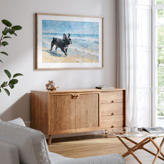 Framed brindle French Bulldog beach watercolor print displayed in a styled interior with soft blue ocean and tan sand tones.