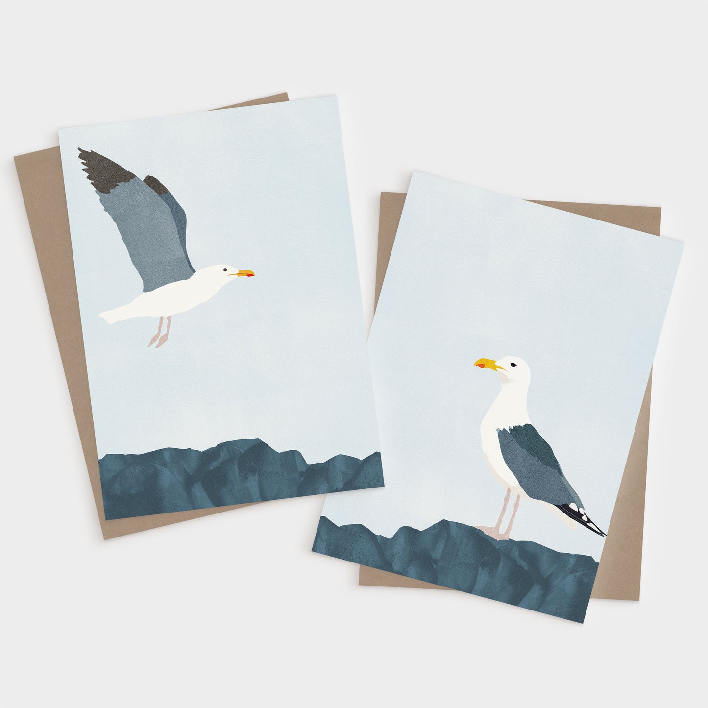 Set of two minimalist greeting cards with white envelopes; one features a seagull in flight above blue rocks, and the other shows a seagull standing on the rocks against a pale sky.