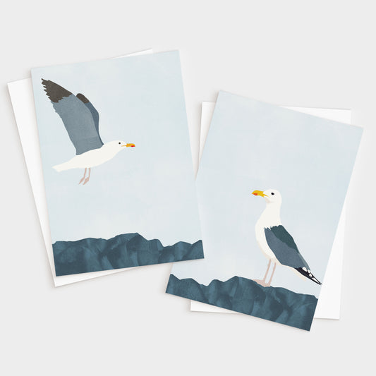 Set of two minimalist greeting cards with white envelopes; one features a seagull in flight above blue rocks, and the other shows a seagull standing on the rocks against a pale sky.