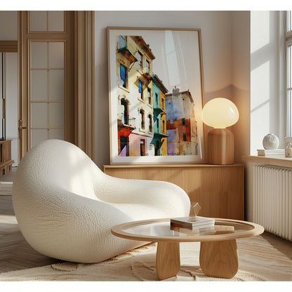 Framed watercolor painting of colorful buildings with pastel facades, leaning against a wall in a sunlit room with a modern chair and wooden table.