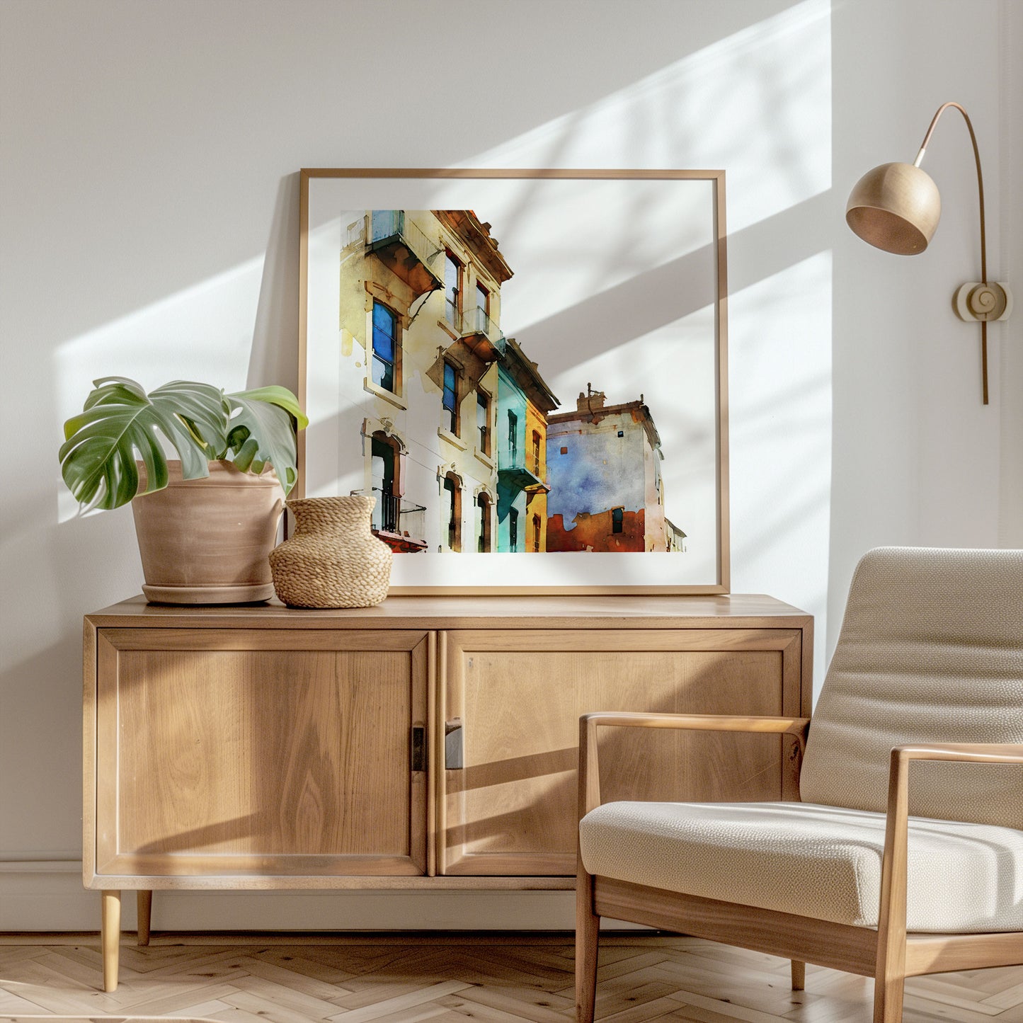 Framed watercolor painting of colorful buildings with pastel facades, placed on a wooden sideboard in a sunlit corner with a plant and chair.
