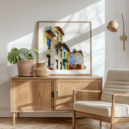 Framed watercolor painting of colorful buildings with pastel facades, placed on a wooden sideboard in a sunlit corner with a plant and chair.