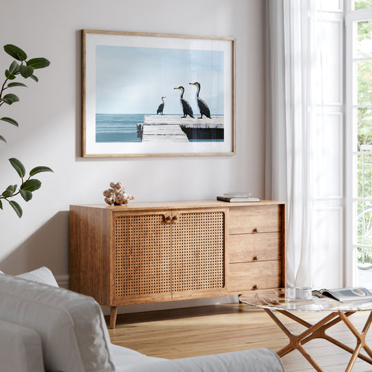 Framed artwork of three cormorants on a pier hangs above a wooden cabinet in a bright, coastal-style living room. The scene features a calm blue sea and sky, with natural light streaming through sheer white curtains.