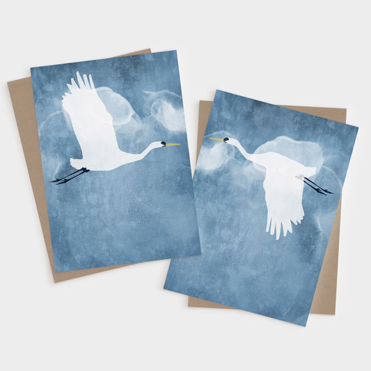 Set of two greeting cards featuring minimalist illustrations of white egrets in flight against a textured blue sky, one with wings raised and the other with wings down, each paired with a kraft brown envelope.