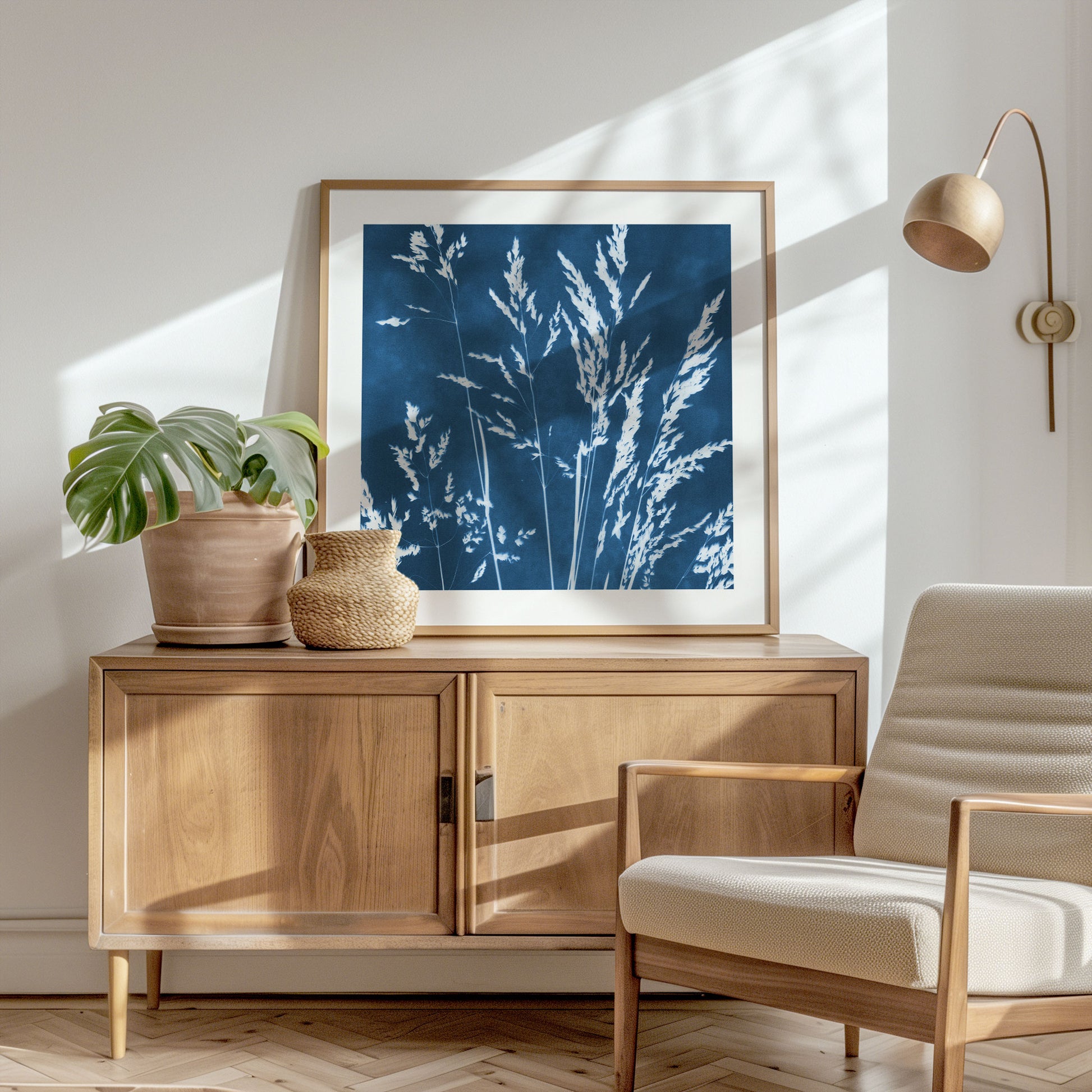 Framed cyanotype print of wild grasses on a wooden sideboard, complemented by a potted monstera, woven vase, and a beige lounge chair in a sunlit room.