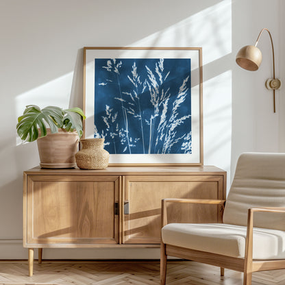 Framed cyanotype print of wild grasses on a wooden sideboard, complemented by a potted monstera, woven vase, and a beige lounge chair in a sunlit room.