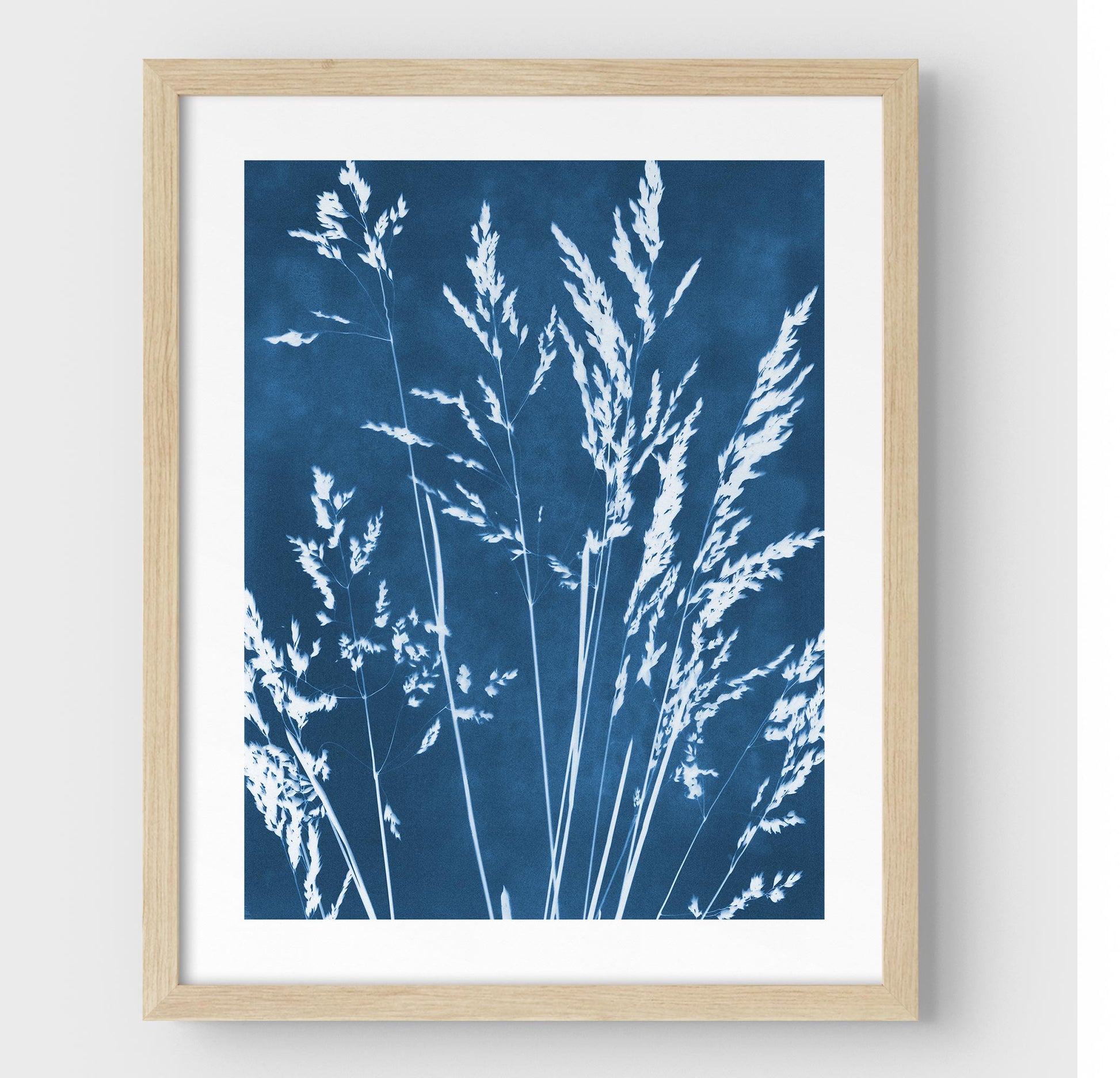 Framed cyanotype print featuring delicate wild grasses in white against a deep blue background, enclosed in a natural wood frame with a white border.