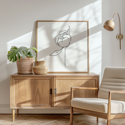 Framed minimalist line drawing of a peony bud with a single leaf, placed on a wooden sideboard in a sunlit modern interior with neutral tones.