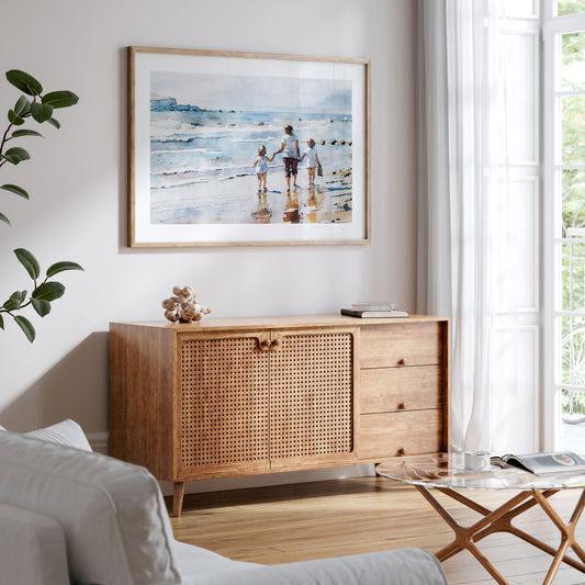 Framed watercolor painting of a woman and two children walking along the shoreline, mounted on a white wall above a wooden sideboard in a bright living space.
