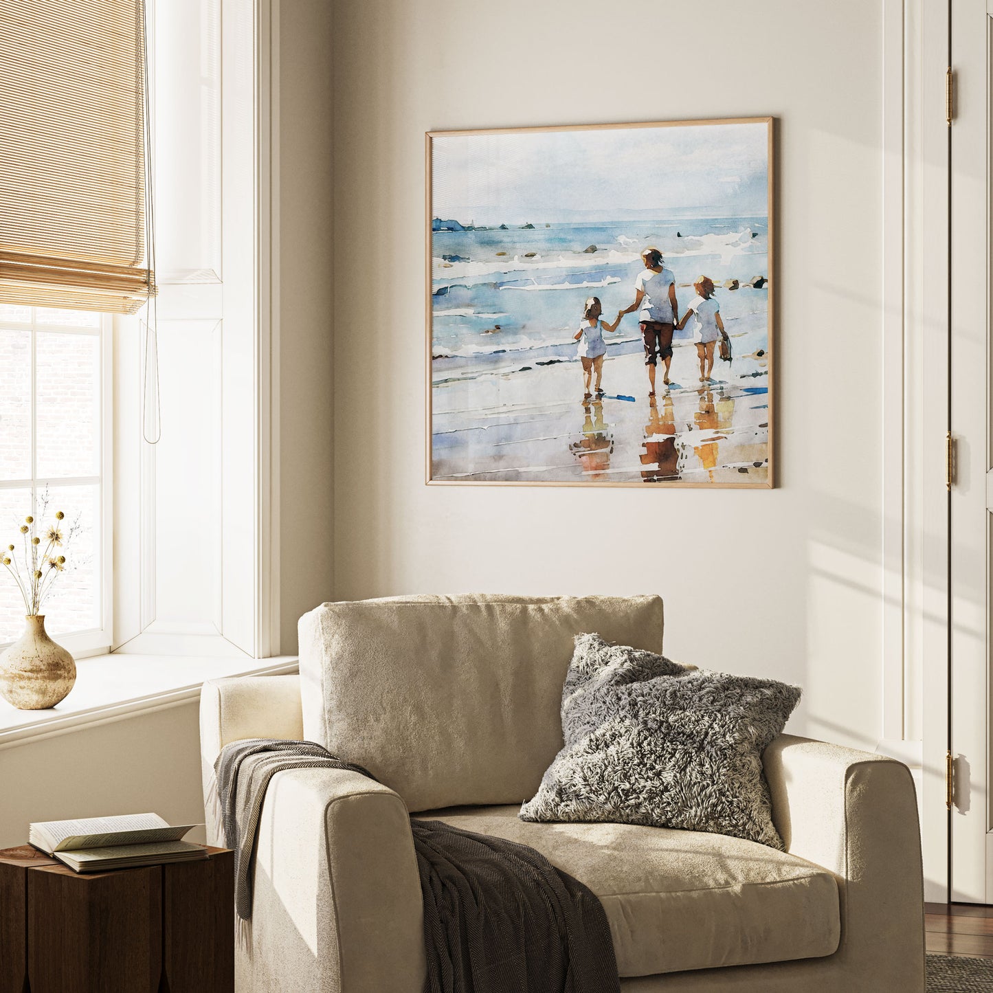 Framed watercolor painting of a woman and two children walking along the shoreline, mounted on a beige wall above a cozy armchair with textured pillows.