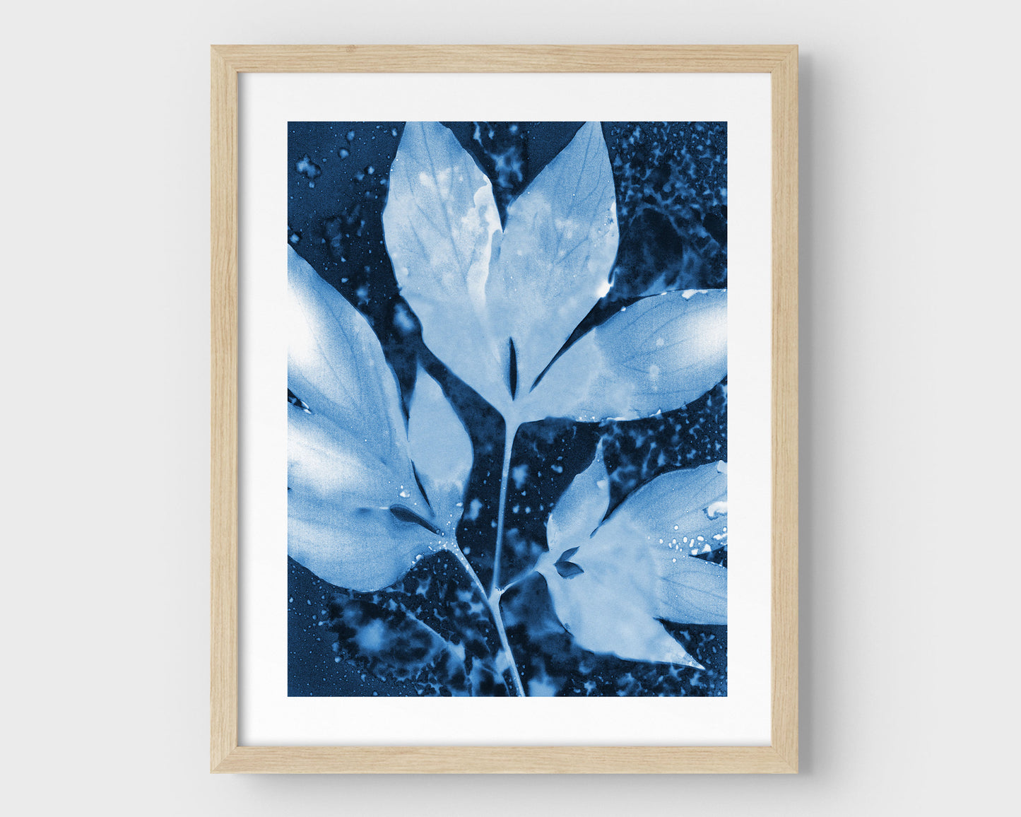 Framed cyanotype print of peony leaves in rich indigo and soft blue tones, featuring delicate leaf silhouettes and organic speckling, set against a clean white background with a natural wood frame.