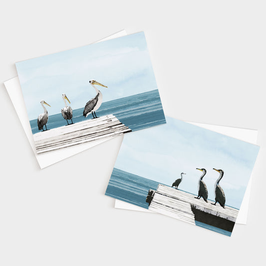 Two illustrated greeting cards featuring coastal birds—one shows three pelicans on a white dock over blue water, and the other shows three cormorants on a similar dock, both under soft blue skies.
