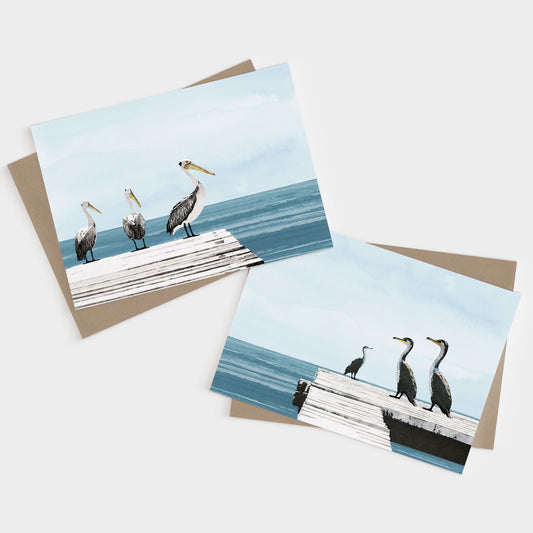 Two illustrated greeting cards featuring coastal birds—one shows three pelicans on a white dock over blue water, and the other shows three cormorants on a similar dock, both under soft blue skies.