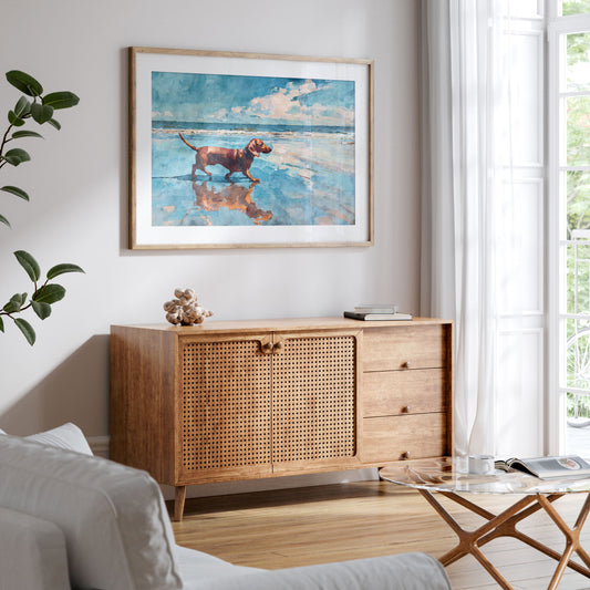 Framed watercolor-style beach scene of a red dachshund walking along the shoreline, reflected in wet sand under a bright blue sky. Displayed above a mid-century wood sideboard with cane doors in a light, airy living room with neutral tones and soft natural light.