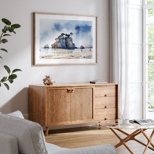 Bright living room with a wooden sideboard, a glass coffee table, and a framed watercolor painting of a coastal rock formation on the wall.